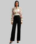 Woman in a floral blouse and black pants standing in a white background.