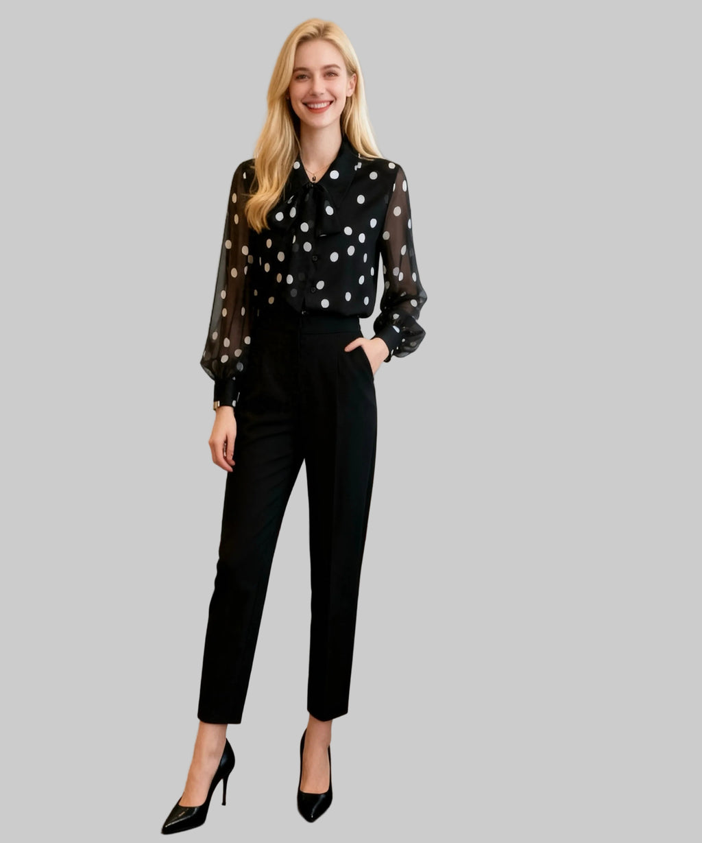 Woman in a polka dot blouse and black pants standing in a white background.