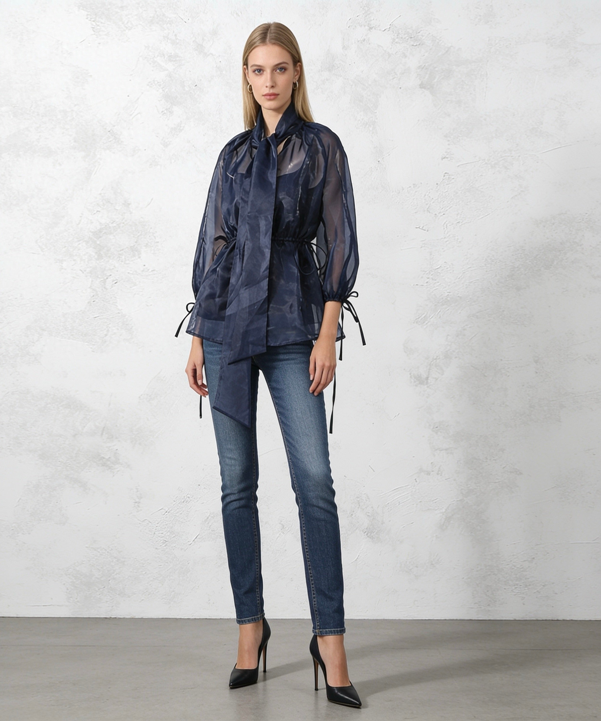 Woman wearing a dark blue outfit with sheer top and jeans against a light gray background