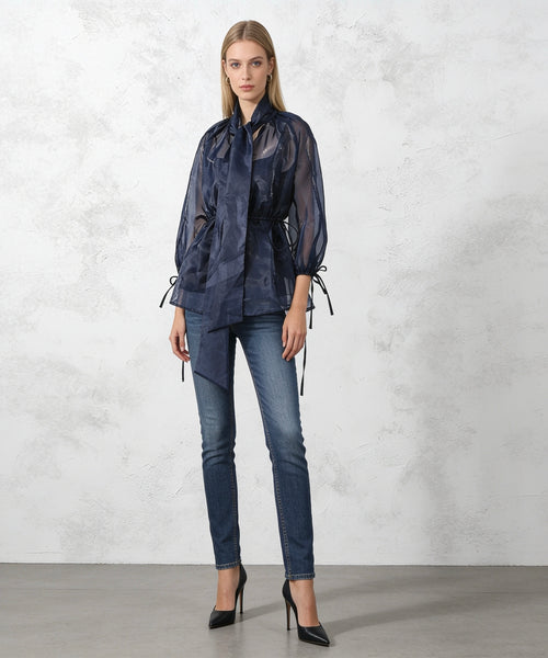 Woman wearing a dark blue outfit with sheer top and jeans against a light gray background