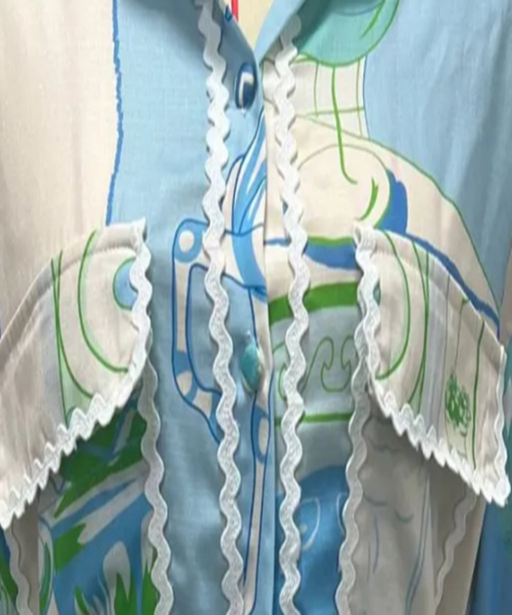 Close-up of a light blue button-up shirt with a green and white graphic print, featuring white scalloped lace trim down the front and on the chest pockets.