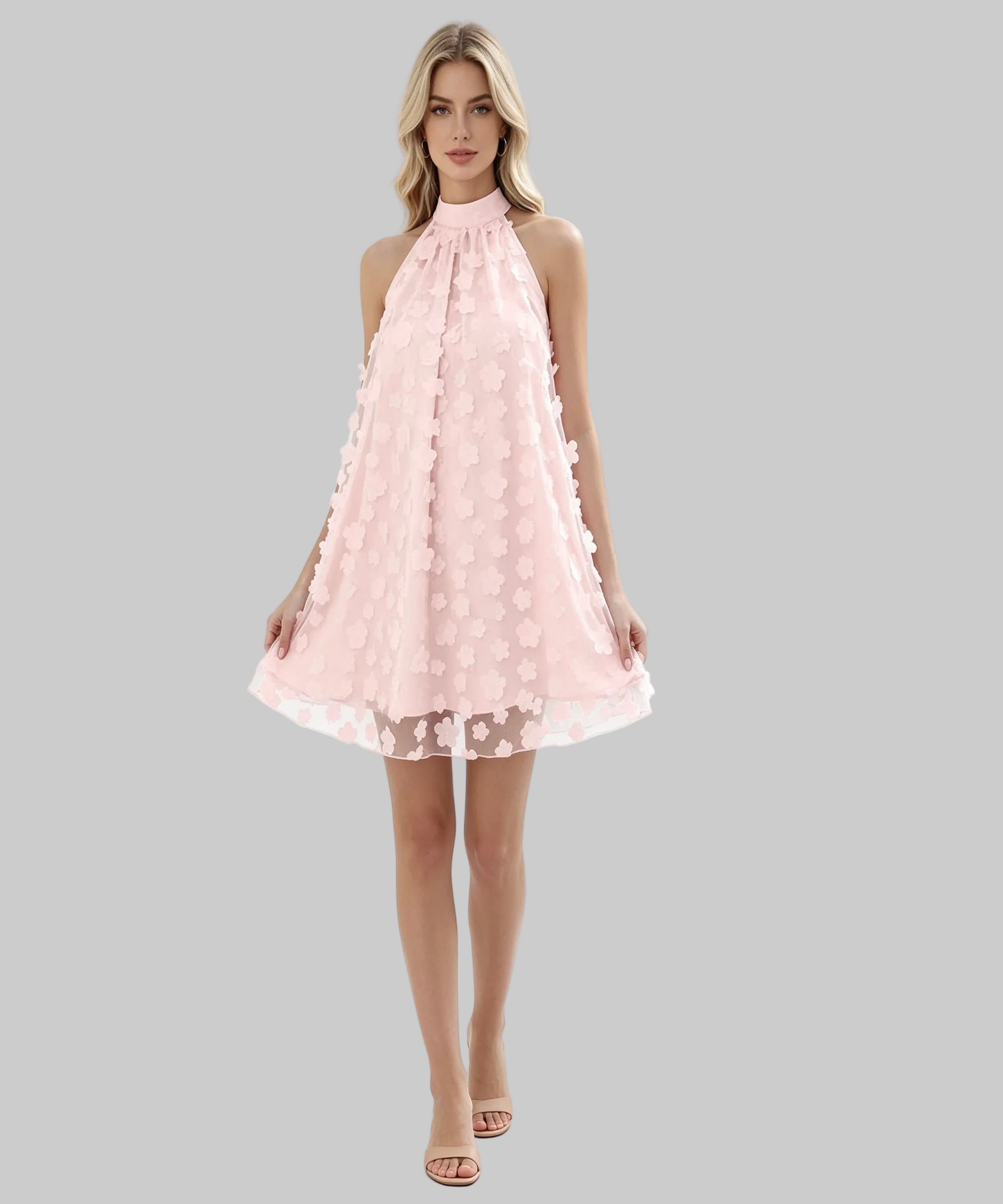 A woman modeling a light pink sleeveless halter dress with a textured, raised floral pattern and a sheer tulle hem, set against a plain white studio background.
