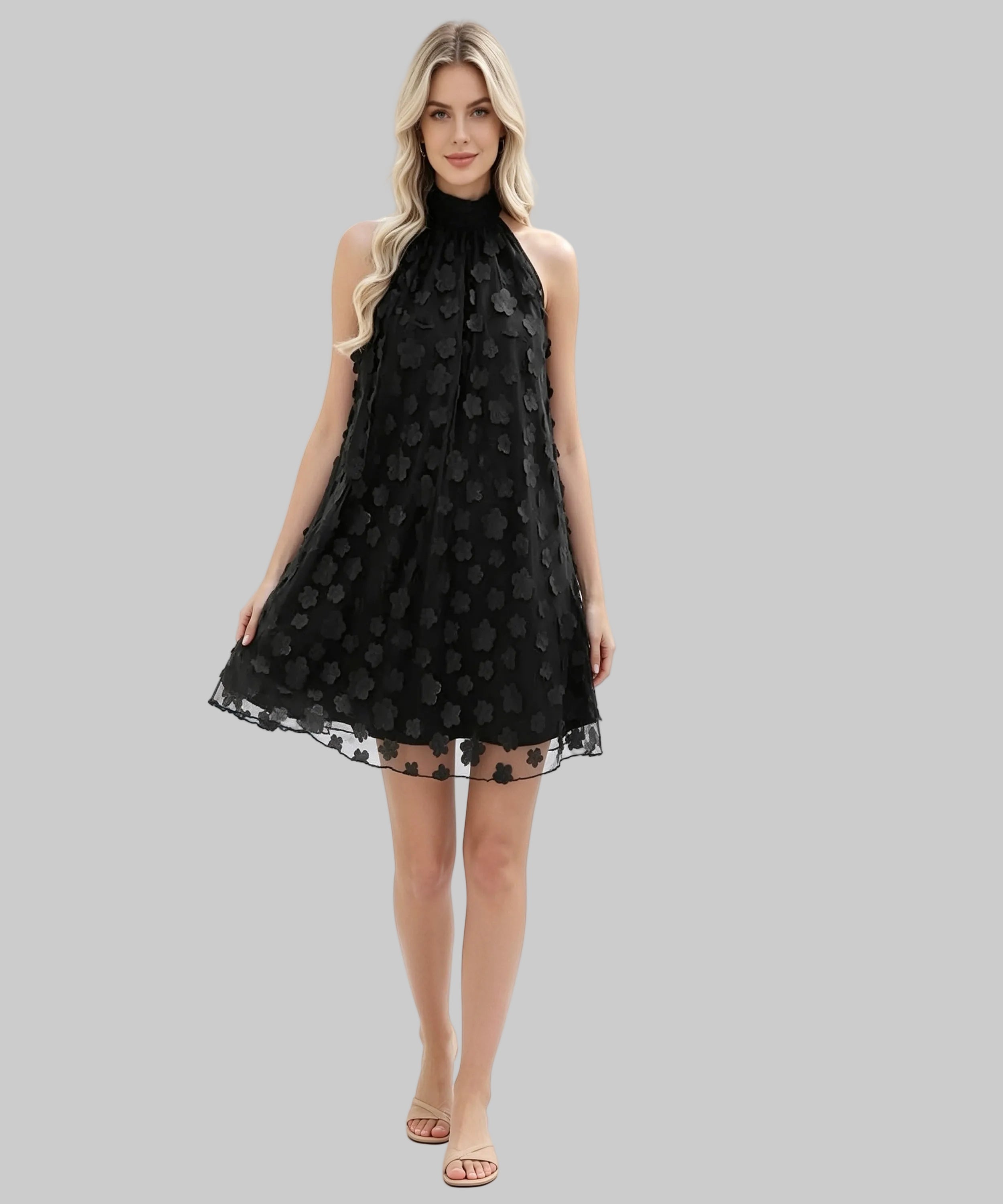 A blonde model wearing a sleeveless black halter-neck mini dress with a textured, raised floral pattern and a sheer tulle hem, standing against a plain white studio background.