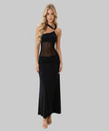 A blonde model wearing a black halter-neck maxi dress with a sheer mesh midsection panel and a draped skirt, paired with beige strappy heels, against a plain white studio background.