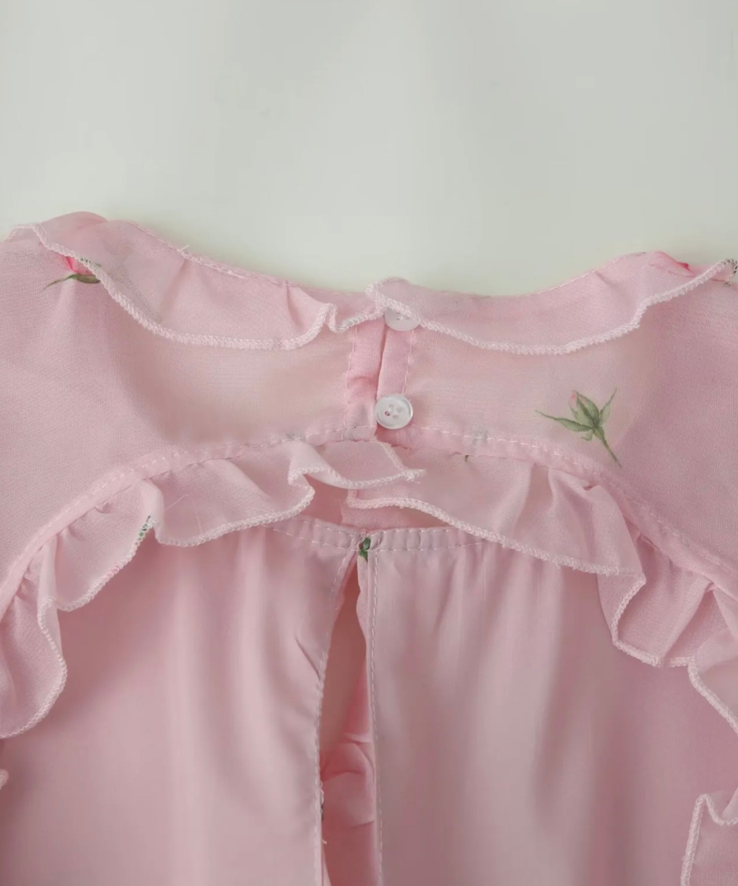 Close-up of a light pink ruffled blouse or nightgown with a button closure and small green floral embroidery, shown against a plain light background.