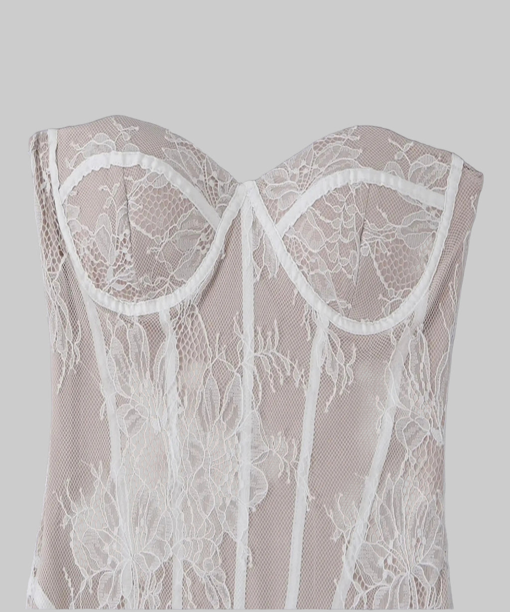 A close-up of a strapless lace corset top in a light beige or taupe color with white floral lace overlays and white trim, laid flat against a neutral gray background.