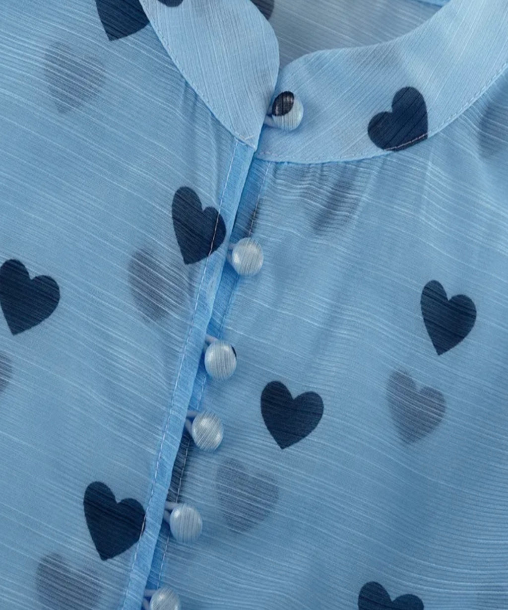 Close-up of a light blue sheer fabric garment with a collar and a pattern of dark blue and gray hearts, featuring a button-down front.