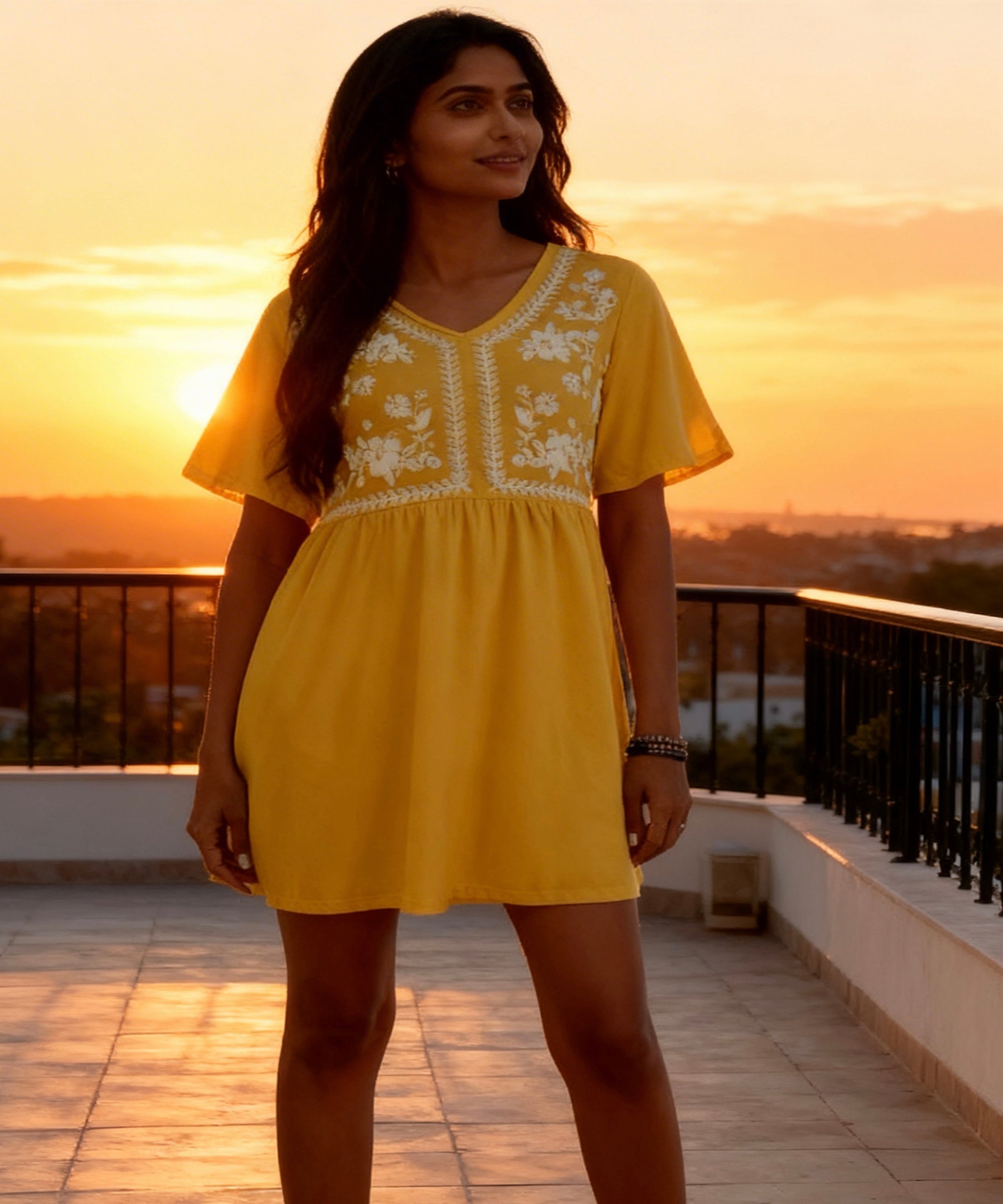 Woman in a yellow dress with floral embroidery standing on a balcony at sunset.
