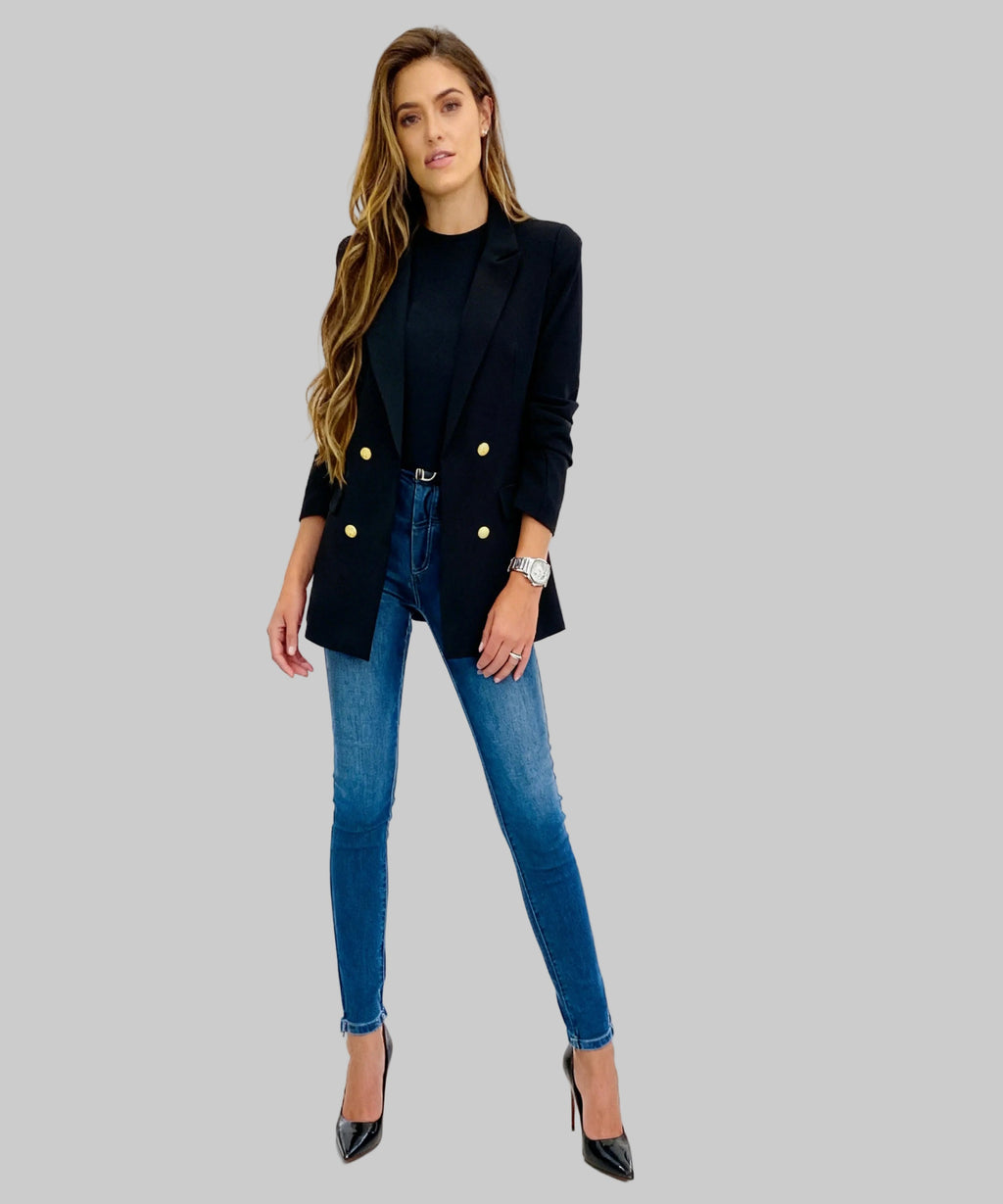 Woman wearing a black blazer with gold buttons and blue jeans on a white background