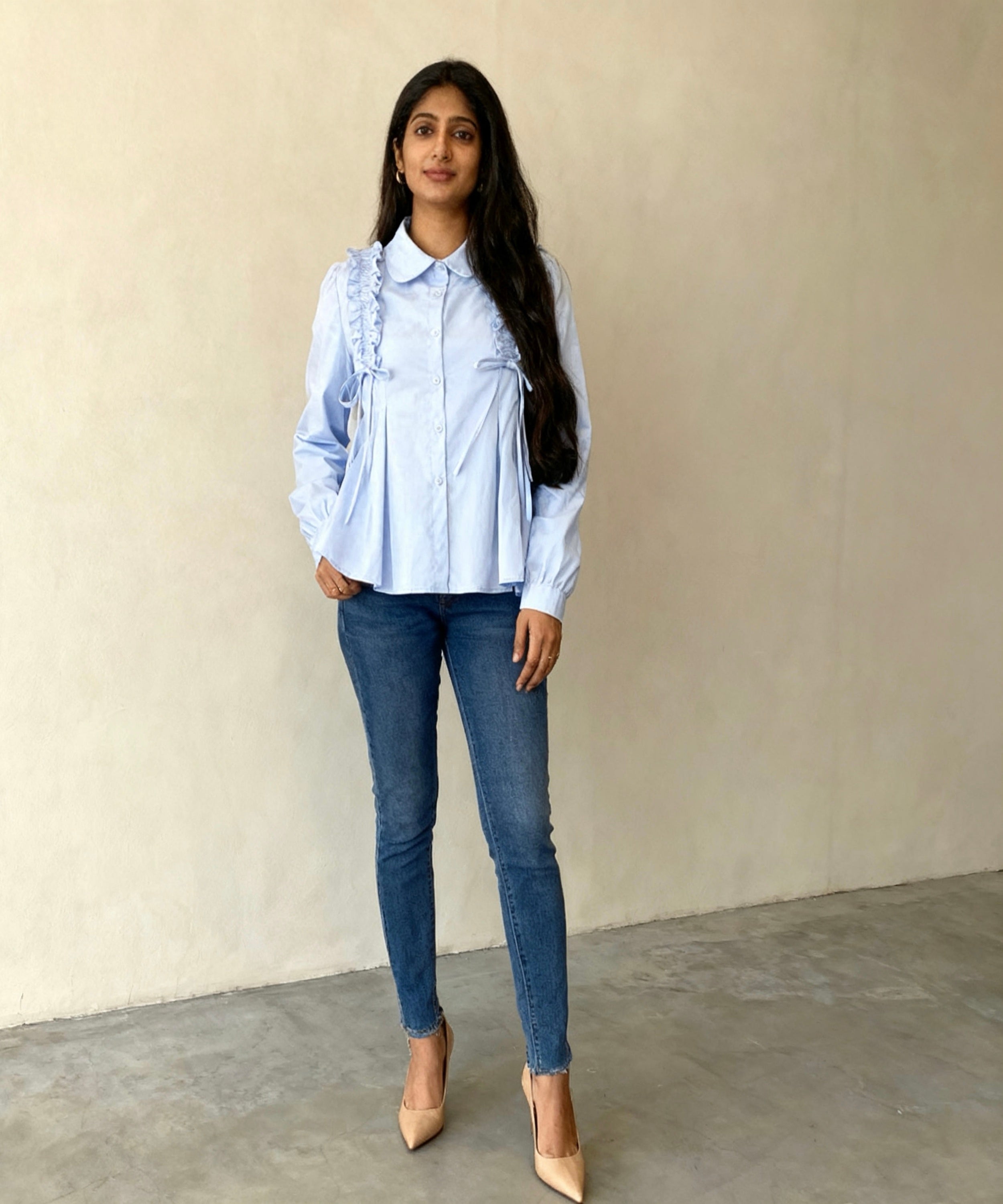 Woman wearing a light blue blouse with ruffles and blue jeans against a beige wall.
