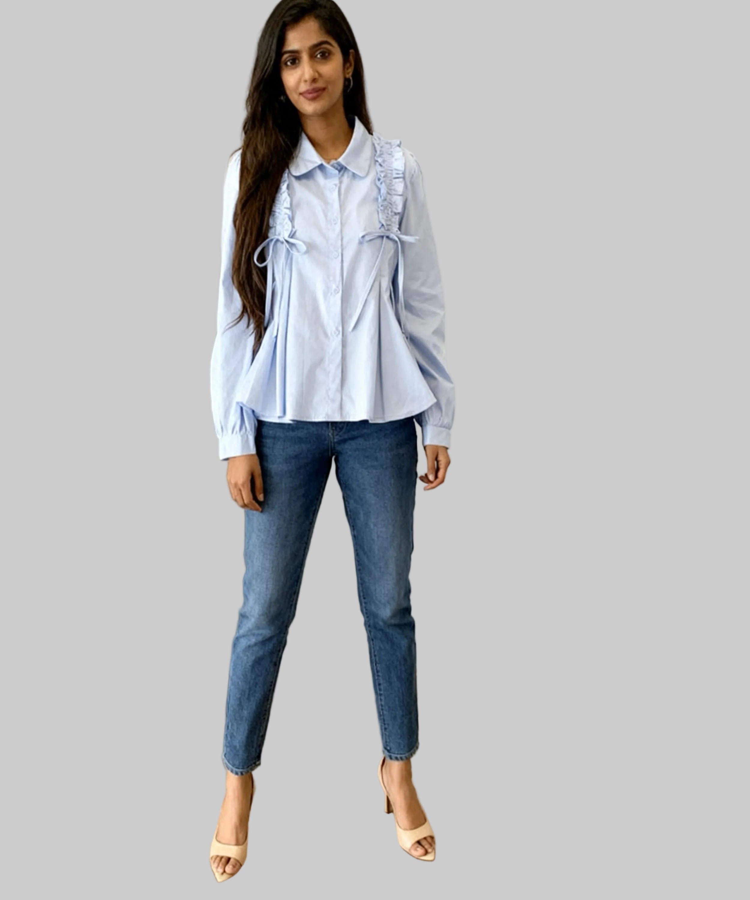 Woman wearing a light blue blouse with lace details and blue jeans standing in a room with a wooden floor and black cabinet.