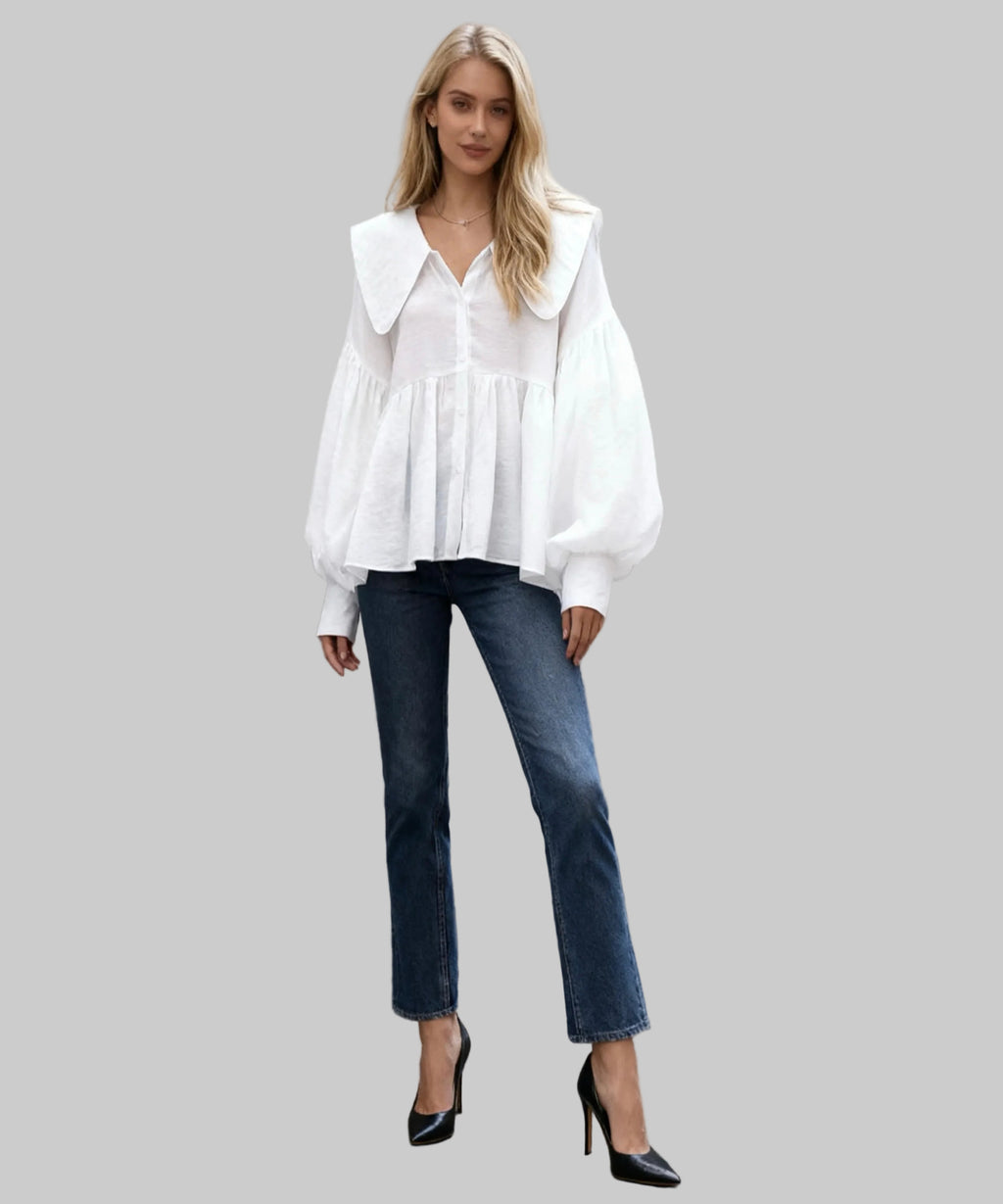 Woman wearing a white blouse and blue jeans standing against a light-colored wall.