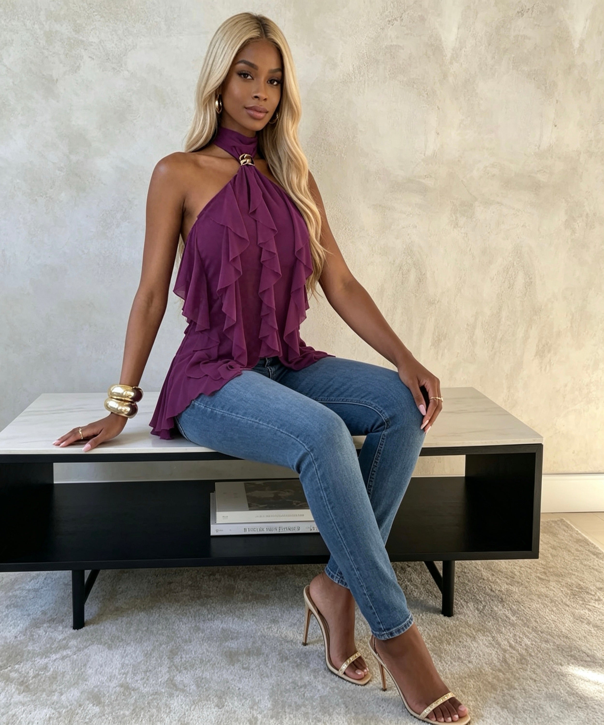Woman in a purple top and blue jeans sitting on a bench against a neutral wall.