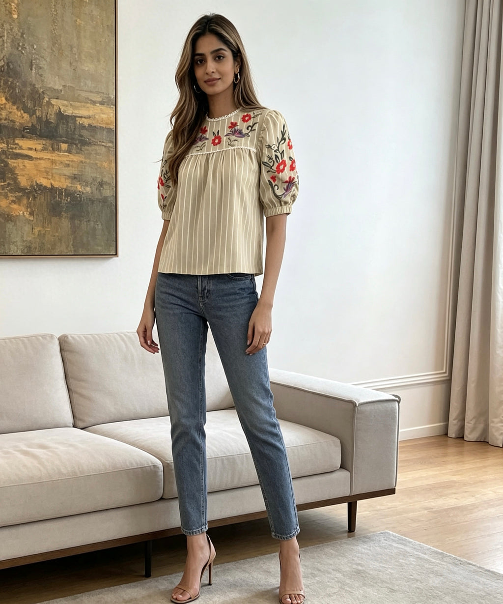 Woman wearing a beige blouse with floral embroidery and blue jeans in a living room.