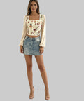 Woman wearing a floral top and denim skirt on a balcony with a cityscape view.