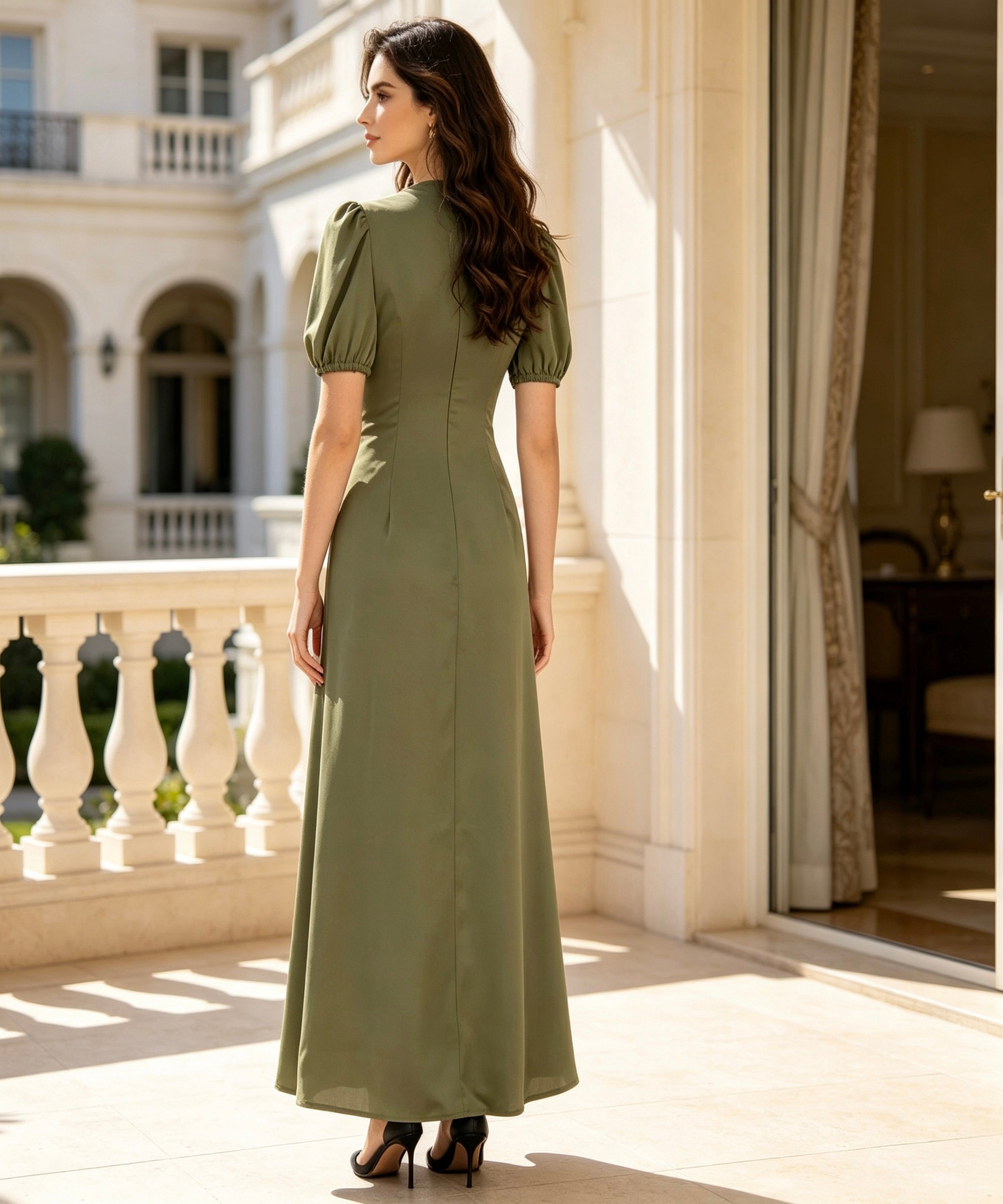 A woman stands on a sunlit balcony wearing a floor-length olive green dress with short puff sleeves, a high neckline, and a fitted silhouette. The photo is taken from behind, showing the back of the dress, with a grand building and windows in the background.
