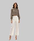 A woman stands on a white background, wearing a taupe cropped long-sleeve blouse with button detail and white wide-leg cropped pants, paired with nude high-heeled sandals.