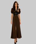 A woman stands on a white background wearing a long brown dress with puff sleeves, a deep V-neckline, and black lace detailing across the bodice. 