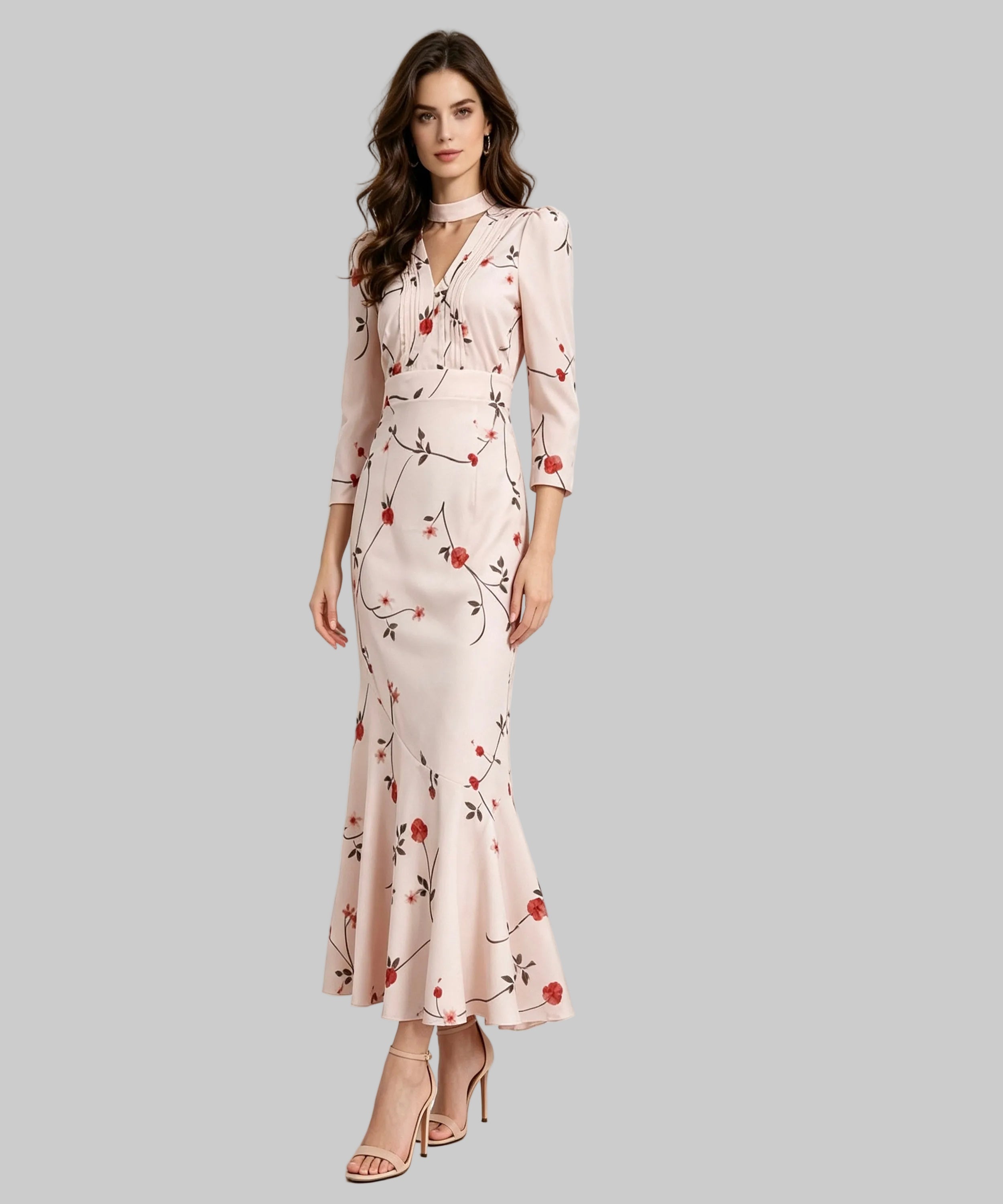 A woman models a long-sleeved, floor-length blush pink dress with a red and green floral print, standing in a minimalist white studio setting.