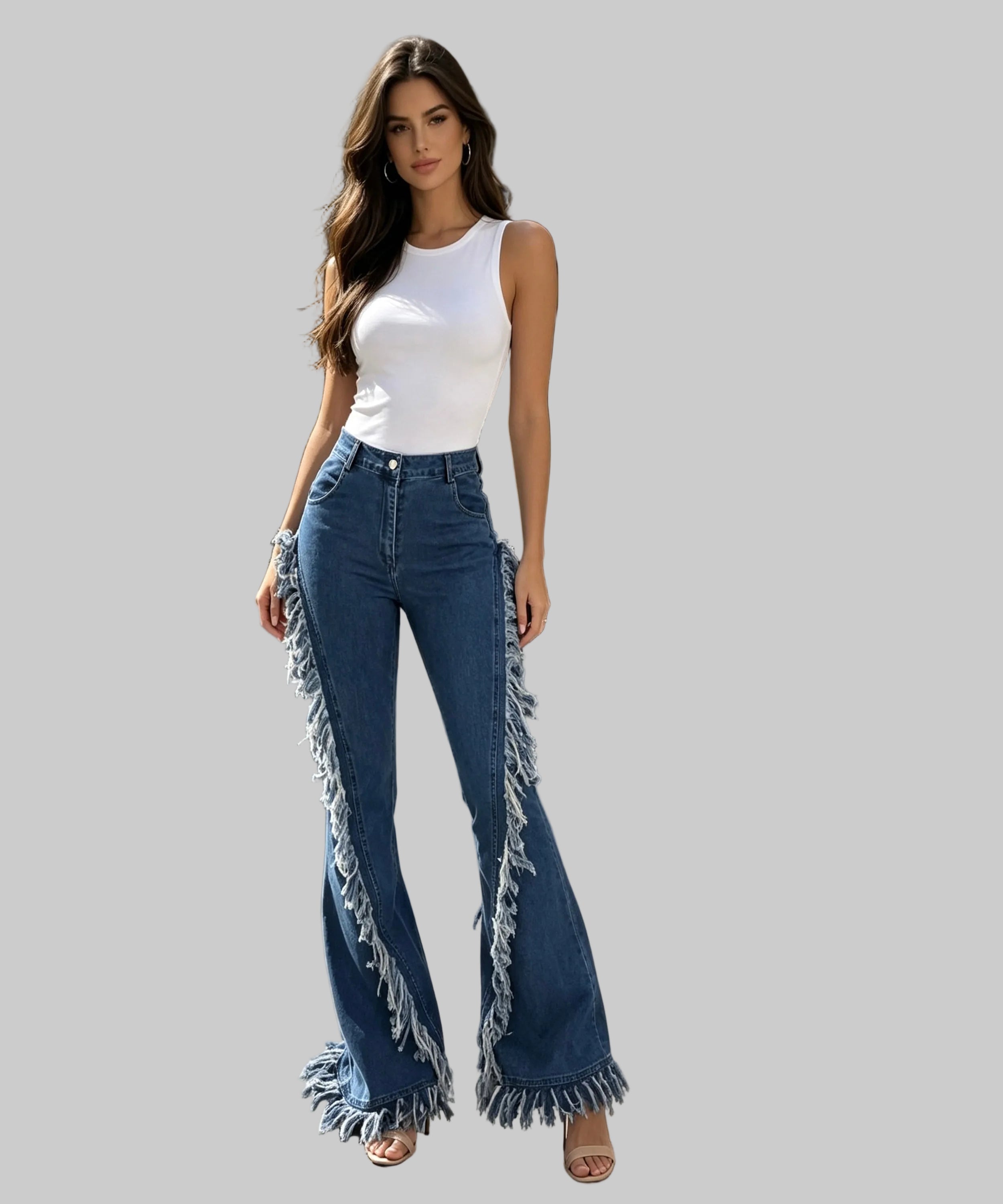 A woman stands on a white background wearing high-waisted blue flare jeans with long fringe detailing running down the outer legs, paired with a white sleeveless top and tan heeled sandals.