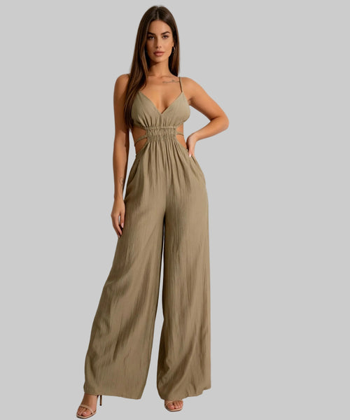 A woman modeling a beige wide-leg jumpsuit with a cutout bodice and thin spaghetti straps, standing against a plain light gray studio background.